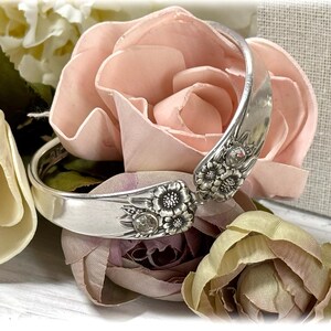 May include: A silver spoon bracelet with floral designs and clear rhinestones. The bracelet is displayed on a pink and white floral background.