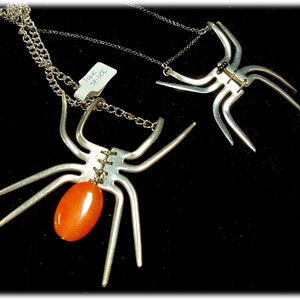 May include: Two silver spider necklaces. One spider has a large orange stone in the centre of its body. The other spider has black beads for eyes. The necklaces are on a black background. The price tag reads "Fork £9.00".