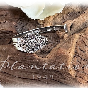 May include: A silver spoon bracelet with a floral pattern on the spoon's bowl. The bracelet is presented on a piece of aged wood. The word "Plantation" and the year "1948" are visible in the image.