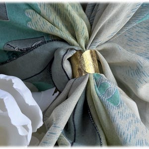 May include: A gold-colored scarf ring holds a patterned scarf in place. The scarf features a design with shades of green, blue, and cream. A white flower is in the foreground. The scarf ring has an etched design.