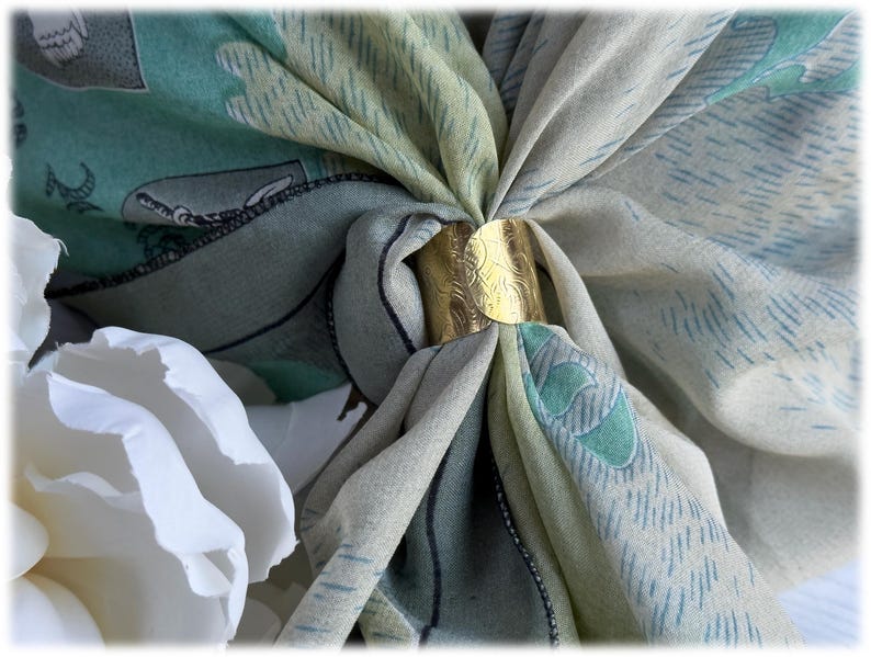 May include: A gold-colored scarf ring holds a patterned scarf in place. The scarf features a design with shades of green, blue, and cream. A white flower is in the foreground. The scarf ring has an etched design.