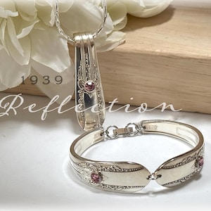 May include: A silver necklace and bracelet set. The necklace pendant and bracelet links are made from repurposed silverware. Each piece features a pink gemstone. The word "Reflection" is written in cursive.