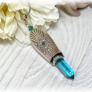 May include: A silver pendant necklace with a detailed floral design and a teal blue crystal point. The pendant is shaped like a teardrop and has a small teal blue gemstone at the top.