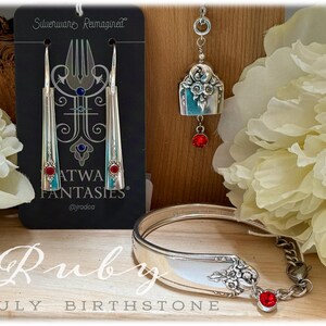 Puede incluir: Juego de joyas "Silverware Reimagined" con pendientes, colgante y pulsera. Los pendientes y la pulsera tienen detalles rojos y azules. El colgante tiene una gema roja. El texto "Ruby July Birthstone" es visible.