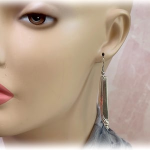May include: A silver-toned earring with a rectangular, elongated design. The earring is suspended from a small hook and features a decorative element at the bottom. The earring is shown on a mannequin.