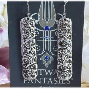May include: Silver rectangular earrings featuring a floral pattern. The earrings are long and narrow, covered in a detailed design of small flowers. They hang from silver hooks. The earrings are displayed against a dark background.
