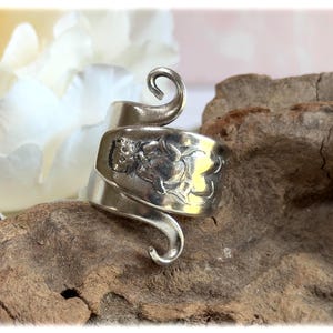 May include: A silver-toned ring with an embossed floral design and a curled, open-ended band. The ring is wrapped around a piece of wood, with a white flower in the background. The ring's design is detailed and intricate.