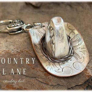 May include: A silver-toned metal cowboy hat charm featuring an embossed floral pattern on the brim, a dark wire band around the crown, and a lobster clasp attachment. The text 1954 COUNTRY LANE cowboy hat is visible on the left side of the image.
