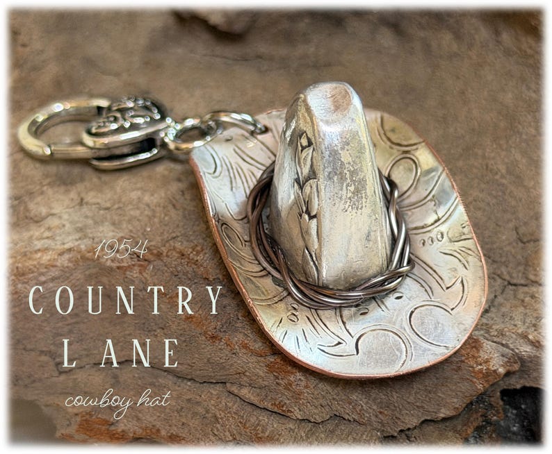 May include: A silver-toned metal cowboy hat charm featuring an embossed floral pattern on the brim, a dark wire band around the crown, and a lobster clasp attachment. The text 1954 COUNTRY LANE cowboy hat is visible on the left side of the image.