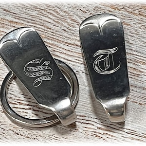 Puede incluir: Dos llaveros de plata con iniciales grabadas, "S" y "T", en una fuente de letra decorativa. Los llaveros están hechos de cubiertos reutilizados.