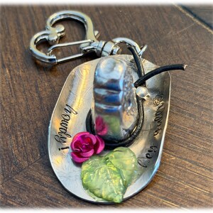 May include: A silver-toned metal keychain shaped like a cowboy hat with a heart-shaped clasp. The hat features the stamped text "howdy!" and "hey y'all", a small pink rose, a translucent green leaf, and a thin black cord wrapped around the crown.