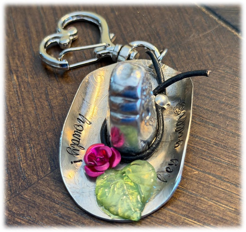 May include: A silver-toned metal keychain shaped like a cowboy hat with a heart-shaped clasp. The hat features the stamped text "howdy!" and "hey y'all", a small pink rose, a translucent green leaf, and a thin black cord wrapped around the crown.