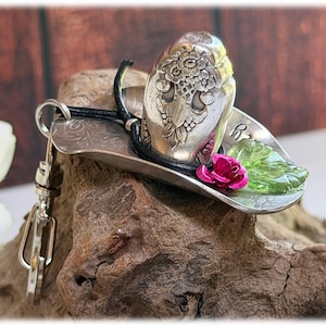 May include: A silver-toned metal keychain shaped like a cowboy hat, featuring an embossed floral pattern on the crown, a black cord band, a small pink rose accent, and a translucent green leaf embellishment, attached to a silver metal lobster clasp.