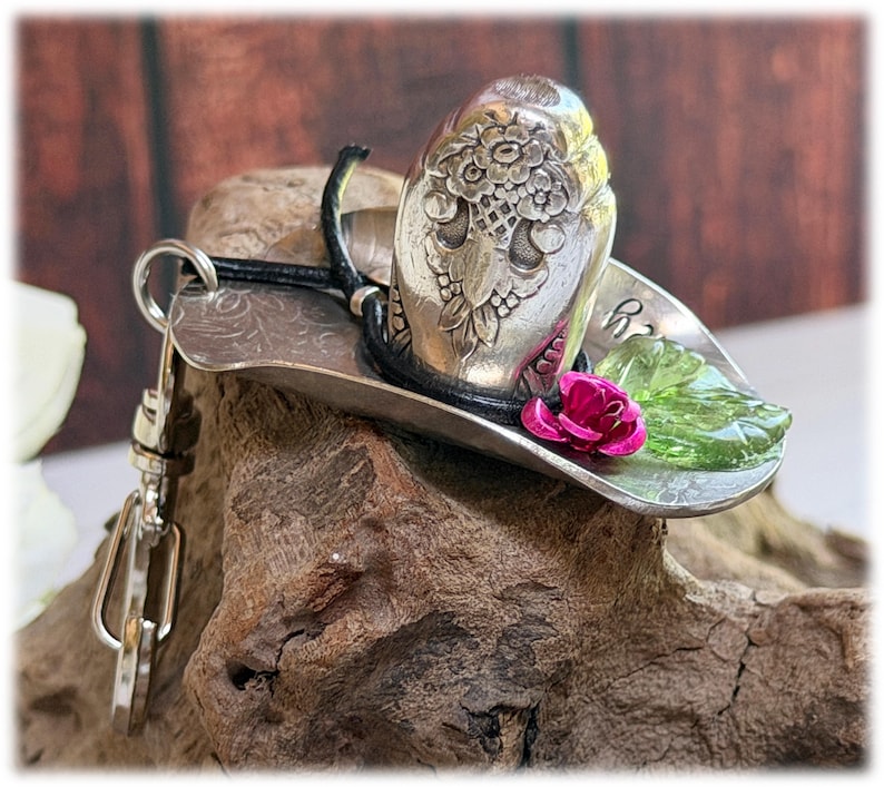 May include: A silver-toned metal keychain shaped like a cowboy hat, featuring an embossed floral pattern on the crown, a black cord band, a small pink rose accent, and a translucent green leaf embellishment, attached to a silver metal lobster clasp.