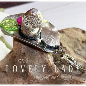 May include: A silver-toned metal keychain shaped like a cowboy hat, featuring an embossed floral pattern, a black cord accent, a small green leaf, a pink flower, and a metal lobster clasp. The text 1937 LOVELY LADY cowgirl hat is visible.