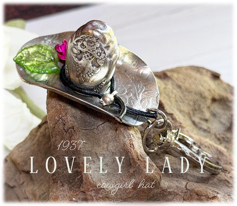 May include: A silver-toned metal keychain shaped like a cowboy hat, featuring an embossed floral pattern, a black cord accent, a small green leaf, a pink flower, and a metal lobster clasp. The text 1937 LOVELY LADY cowgirl hat is visible.