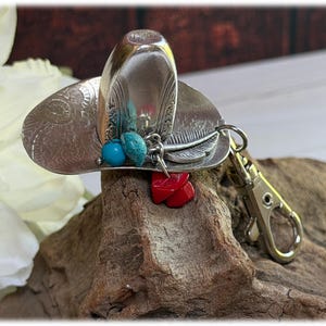 May include: A silver-toned metal keychain shaped like a cowboy hat, featuring an embossed floral pattern on the brim. The hat is adorned with a dangling charm set consisting of a silver feather, two small turquoise-colored beads, and red stones.