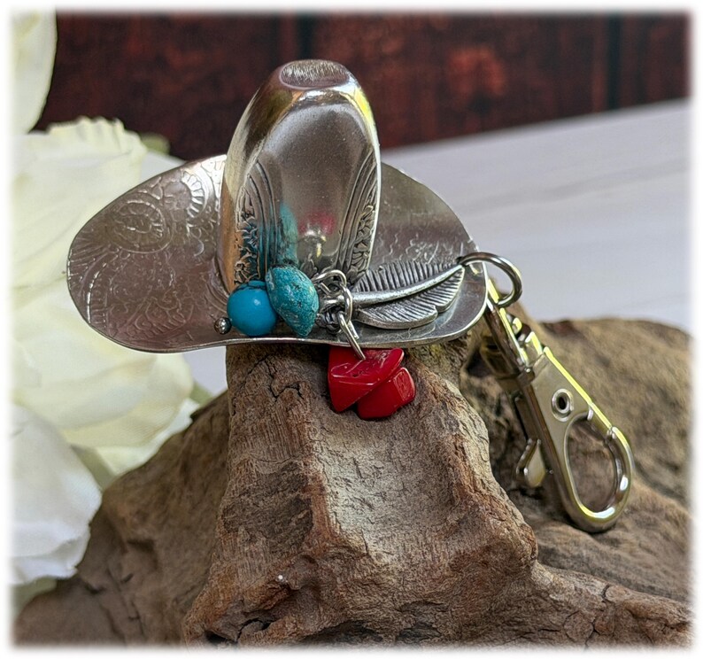 May include: A silver-toned metal keychain shaped like a cowboy hat, featuring an embossed floral pattern on the brim. The hat is adorned with a dangling charm set consisting of a silver feather, two small turquoise-colored beads, and red stones.