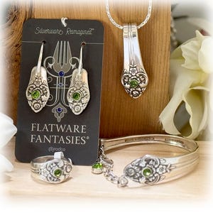 Puede incluir: Una colección de joyas de cubiertos reciclados, que incluye pendientes, un collar con colgante, un anillo y una pulsera. Cada pieza presenta un diseño floral y una piedra preciosa verde. Las joyas se exhiben sobre una superficie de madera con una etiqueta negra "FLATWARE FANTASIES®".