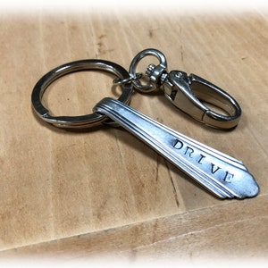 May include: Silver spoon keyring with a ring and a clasp. The spoon has the word "DRIVE" engraved on it.