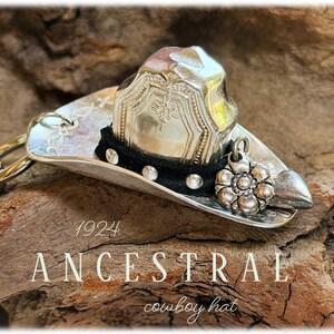 May include: A silver-toned cowboy hat ornament crafted from a vintage spoon, featuring a black band with small silver studs, a floral charm, and a heart-shaped accent. The text 1924 ANCESTRAL cowboy hat is visible on the surface of the object.