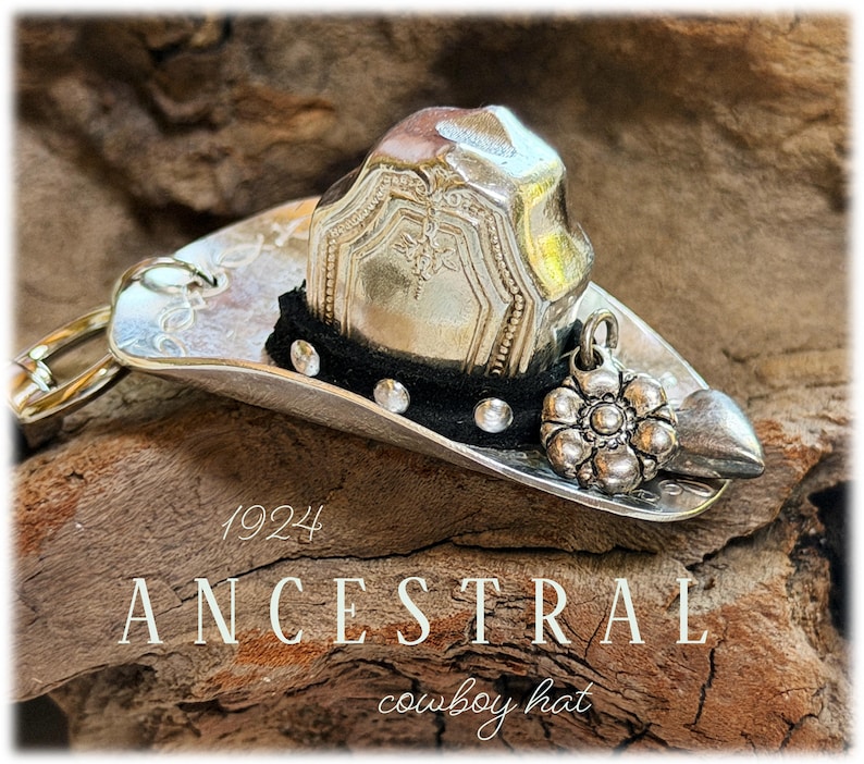 May include: A silver-toned cowboy hat ornament crafted from a vintage spoon, featuring a black band with small silver studs, a floral charm, and a heart-shaped accent. The text 1924 ANCESTRAL cowboy hat is visible on the surface of the object.