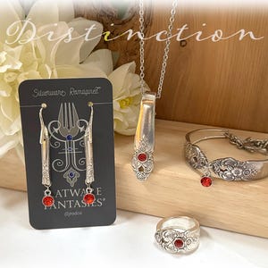 Puede incluir: Una colección de joyas artesanales de plata reciclada, que incluye pendientes, un collar, una pulsera y un anillo. Cada pieza está hecha de cubiertos reciclados y presenta detalles de piedras preciosas rojas. Los pendientes se exhiben en una tarjeta negra con el texto "Silverware Reimagined".