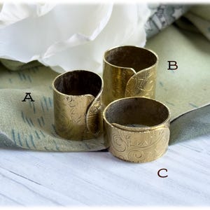 May include: Three brass napkin rings with an embossed floral pattern. The rings are cylindrical with a curved, overlapping design. The rings are different sizes, with the largest being approximately 2 inches tall and the smallest being about 1 inch tall.