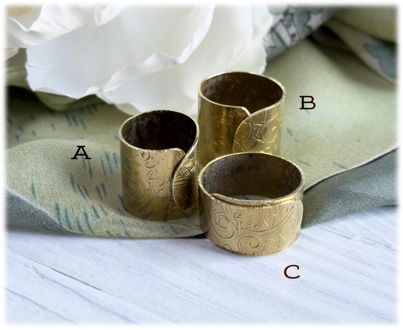 May include: Three brass napkin rings with an embossed floral pattern. The rings are cylindrical with a curved, overlapping design. The rings are different sizes, with the largest being approximately 2 inches tall and the smallest being about 1 inch tall.