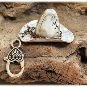 May include: A silver-toned metal cowboy hat charm featuring an embossed floral design on the crown and a copper-colored rim. A dark twisted wire band encircles the base of the crown, and a heart-shaped lobster clasp is attached by a jump ring.