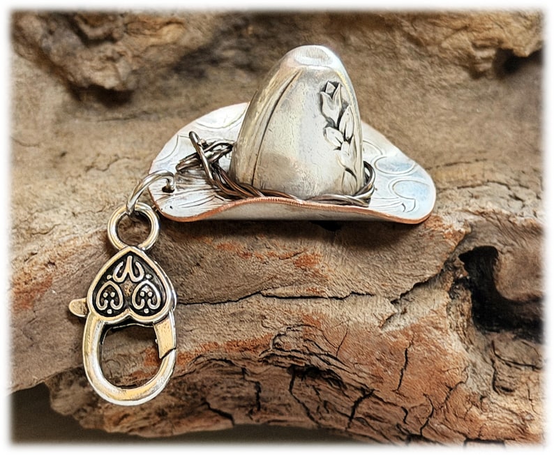 May include: A silver-toned metal cowboy hat charm featuring an embossed floral design on the crown and a copper-colored rim. A dark twisted wire band encircles the base of the crown, and a heart-shaped lobster clasp is attached by a jump ring.