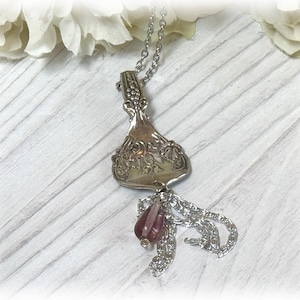 Puede incluir: Un collar con colgante plateado hecho de una cuchara reutilizada. El mango de la cuchara presenta detalles ornamentados, y el colgante está adornado con un grupo de pequeñas cuentas de color malva en forma de lágrima. El collar cuelga de una delicada cadena.