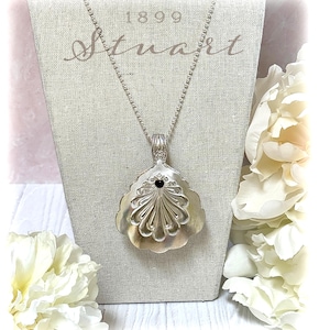 Pode incluir: Colar com pingente prateado com um pingente decorativo em forma de leque. O pingente tem uma pedra preta no centro e é suspenso por uma corrente delicada. A palavra "Stuart" está escrita em itálico acima do pingente.
