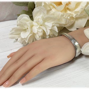 May include: A silver cuff bracelet with an etched design. The bracelet is wide and has a slightly textured surface. The image shows the bracelet on a person's wrist, with white flowers in the background.
