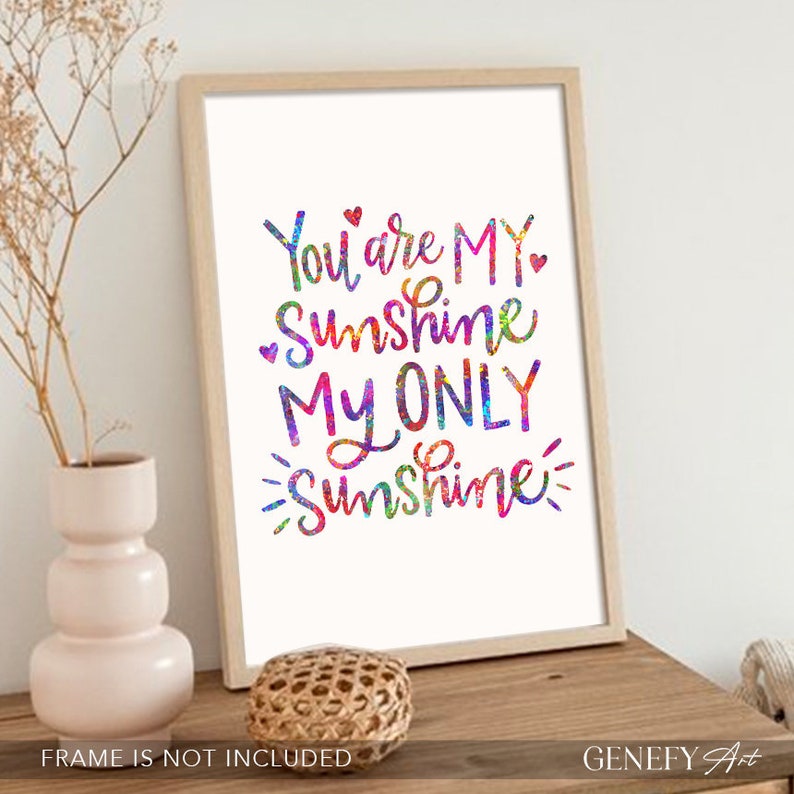 May include: A framed print with the words "You are MY Sunshine MY ONLY Sunshine" in colorful, watercolor-style lettering. The artwork is displayed on a wooden surface, with a decorative vase and woven ball nearby.