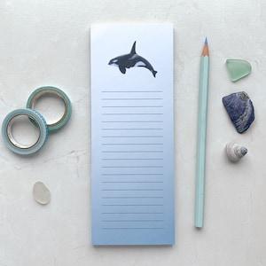 May include: A notepad with a blue gradient background and a black and white orca illustration. Two rolls of decorative washi tape, a light blue pencil, and sea shells are arranged around the notepad. The notepad has horizontal lines for writing.