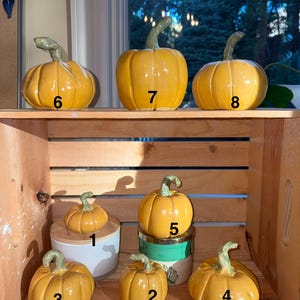 May include: Eight yellow ceramic pumpkins with green stems, numbered one through eight. The pumpkins vary in size and are arranged on two levels within a wooden crate. Each pumpkin features a glossy finish and distinct vertical ridges.