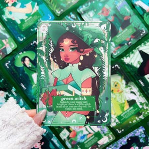 Peut inclure: Illustration d'une sorci&egrave;re verte sur une carte, repr&eacute;sentant une femme aux cheveux fonc&eacute;s, un chapeau vert et une cape. La carte contient le texte "green witch" et une citation sur l'intuition. D'autres cartes avec des illustrations similaires sont en arri&egrave;re-plan.