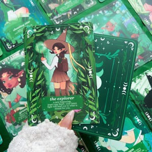 Peut inclure: Un jeu de cartes de tarot avec un th&egrave;me vert et blanc. La carte principale pr&eacute;sente une illustration d'un personnage avec un chapeau de sorci&egrave;re et une jupe, intitul&eacute;e "l'explorateur". D'autres cartes montrent des illustrations et du texte similaires. Les cartes sont entour&eacute;es de feuillage vert.