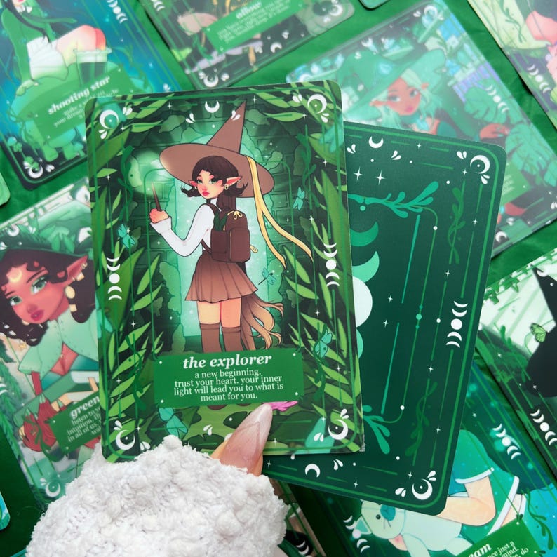 Peut inclure: Un jeu de cartes de tarot avec un th&egrave;me vert et blanc. La carte principale pr&eacute;sente une illustration d'un personnage avec un chapeau de sorci&egrave;re et une jupe, intitul&eacute;e "l'explorateur". D'autres cartes montrent des illustrations et du texte similaires. Les cartes sont entour&eacute;es de feuillage vert.