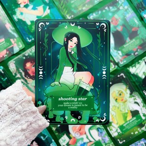 Peut inclure: Une carte de tarot verte avec une illustration d'une personne aux cheveux noirs, v&ecirc;tue d'une tenue verte et assise sur un champignon. La carte porte l'inscription "shooting star" et "make a wish! 111 your dream is meant to be a reality."