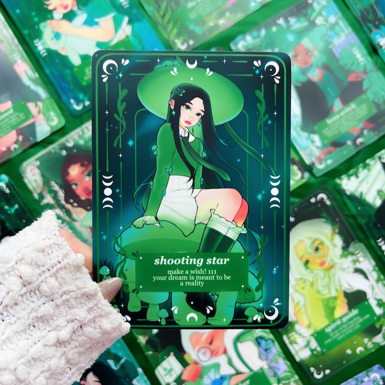 Peut inclure: Une carte de tarot verte avec une illustration d'une personne aux cheveux noirs, v&ecirc;tue d'une tenue verte et assise sur un champignon. La carte porte l'inscription "shooting star" et "make a wish! 111 your dream is meant to be a reality."