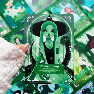 Peut inclure: Une carte de tarot verte avec une illustration de sorci&egrave;re aux longs cheveux verts et un chapeau noir. Un serpent noir l'enlace. Le texte de la carte indique "divine timing" et "trust the unseen, what you're searching for is searching for you."