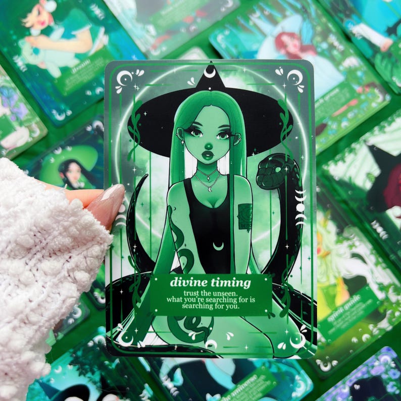 Peut inclure: Une carte de tarot verte avec une illustration de sorci&egrave;re aux longs cheveux verts et un chapeau noir. Un serpent noir l'enlace. Le texte de la carte indique "divine timing" et "trust the unseen, what you're searching for is searching for you."