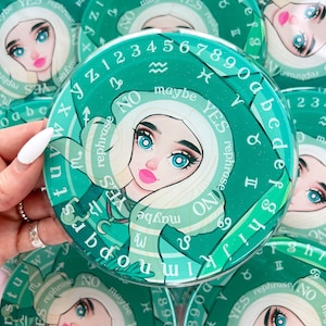 May include: A green and white circular divination board with a woman's face in the center. The board has astrological symbols, numbers, and the words "YES", "NO", "maybe", and "rephrase".