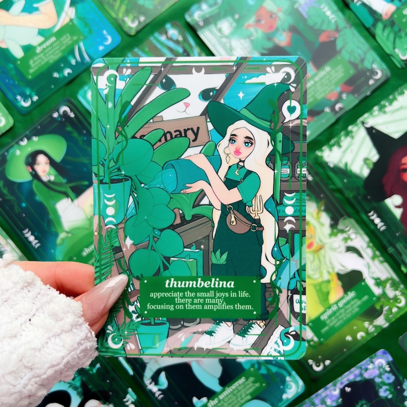 Peut inclure: Une carte illustr&eacute;e verte et blanche repr&eacute;sentant une femme avec un chapeau de sorci&egrave;re arrosant des plantes. La carte contient le texte "thumbelina" et une citation. D'autres cartes avec des illustrations similaires sont en arri&egrave;re-plan.
