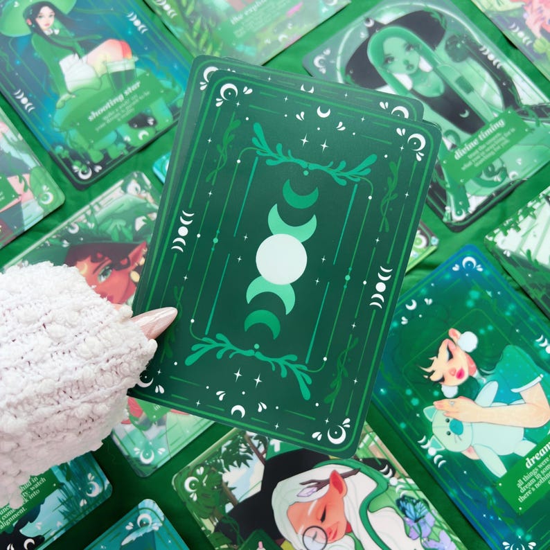 Peut inclure: Un ensemble de cartes de tarot vertes avec des illustrations et du texte. Les cartes pr&eacute;sentent un motif avec des croissants de lune, des &eacute;toiles et des &eacute;l&eacute;ments botaniques. Les cartes sont tenues par une personne portant un pull blanc.