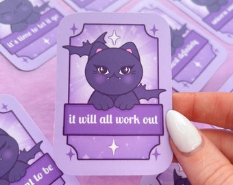 May include: Purple and white sticker with a cartoon black cat with bat wings. The cat is looking at the viewer with a mischievous expression. The sticker has a gold border and the text "it will all work out" in white.
