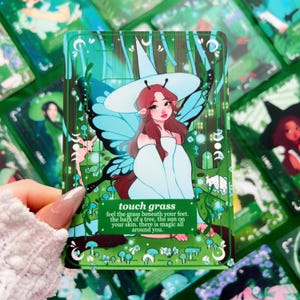 Peut inclure: Une carte d'oracle verte avec une illustration d'une femme avec des ailes de papillon et un chapeau de sorci&egrave;re. La carte indique "touch grass" avec une description po&eacute;tique. D'autres cartes sont visibles en arri&egrave;re-plan.