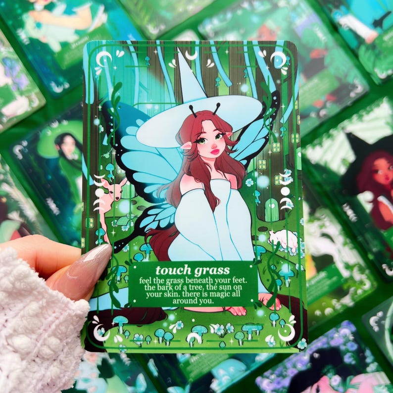Peut inclure: Une carte d'oracle verte avec une illustration d'une femme avec des ailes de papillon et un chapeau de sorci&egrave;re. La carte indique "touch grass" avec une description po&eacute;tique. D'autres cartes sont visibles en arri&egrave;re-plan.